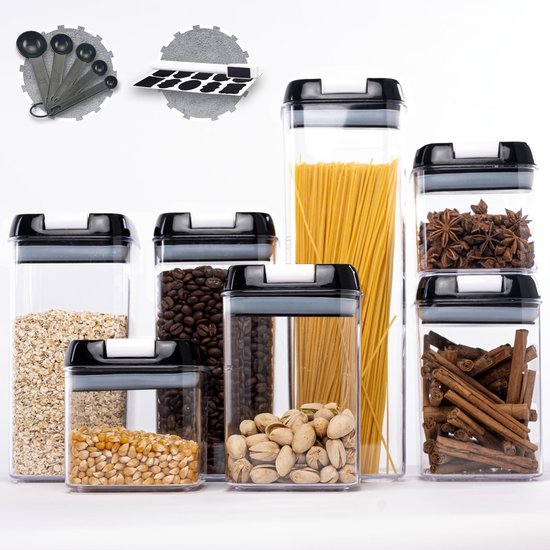 Set van 7 doorzichtige voorraadpotten met zwarte deksels, gevuld met havermout, koffiebonen, pistachenoten, maiskorrels, spaghetti, steranijs en kaneelstokjes. Maatlepels en labels zichtbaar.