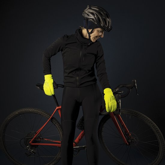 Een vrouw met een zwarte fietsoutfit, helm en neongele handschoenen, lacht naast een rode racefiets.