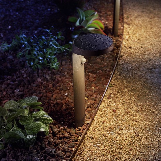 Een natte antracietkleurige sokkellamp met sensor in een tuin met stenen en planten, in het donker.