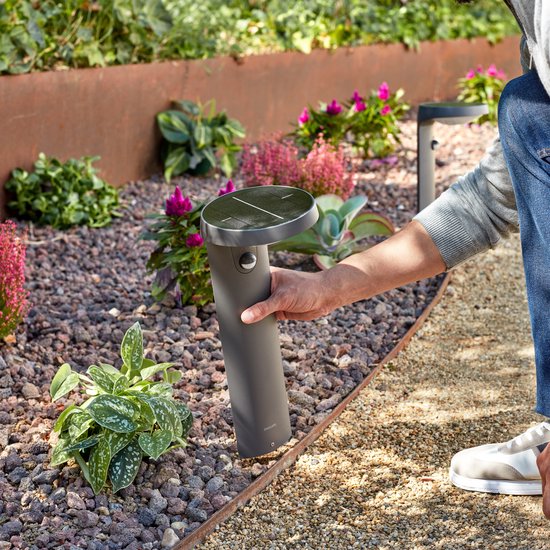 Een hand plaatst een donkere sokkellamp met zonnepaneel in een tuin.