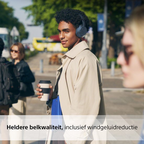 Een man in een beige jas draagt een blauwe Sony WH-CH720N koptelefoon in de stad, met een koffiebeker.