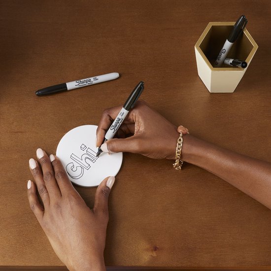 Een vrouw met een armband schrijft "chi" met een Sharpie marker op een witte, ronde onderzetter.