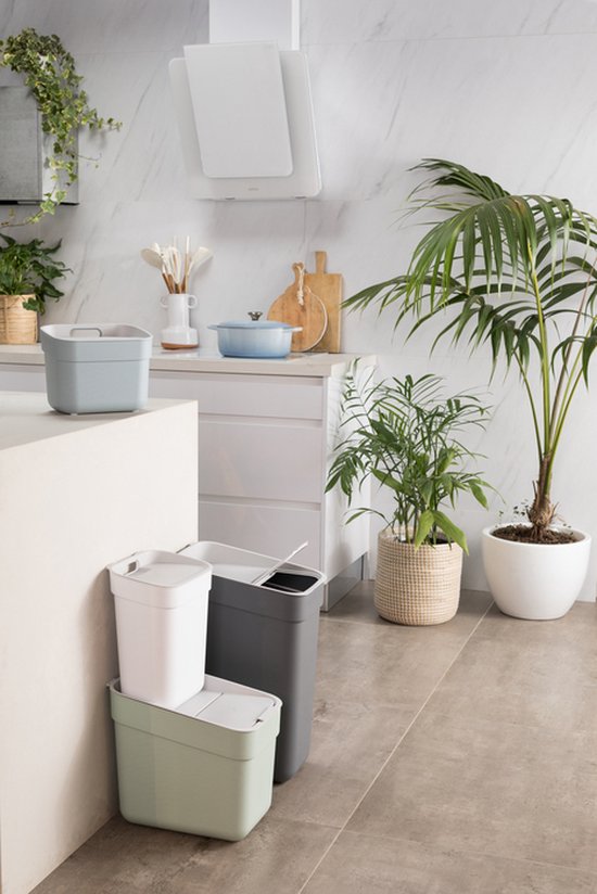 Une poubelle grise, une blanche et une vert clair empilées dans une cuisine. À côté d'une plante.
