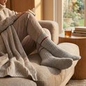 Vrouw met een beige gebreide outfit en grijze sokken met rode streep leest een boek.