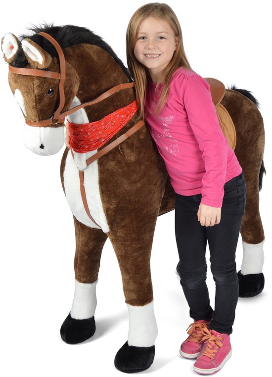 Meisje in roze shirt en zwarte broek naast een groot bruin pluche paard met zadel en rode bandana.