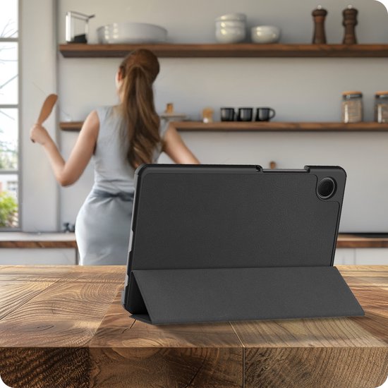 Étui pour tablette noir sur une table en bois, avec une femme cuisinant à l'arrière-plan.