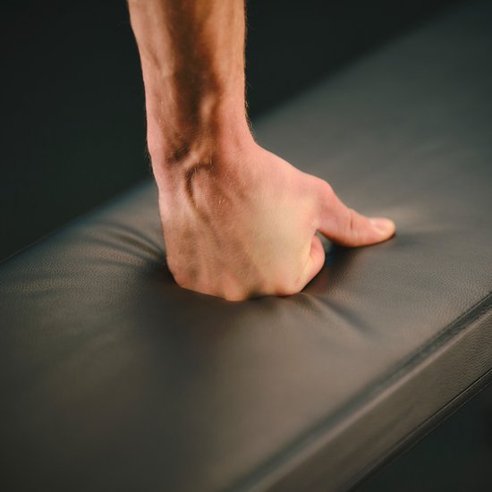 Gedetailleerde close-up van een hand die op de leren zitting van een zwarte trainingsbank drukt.