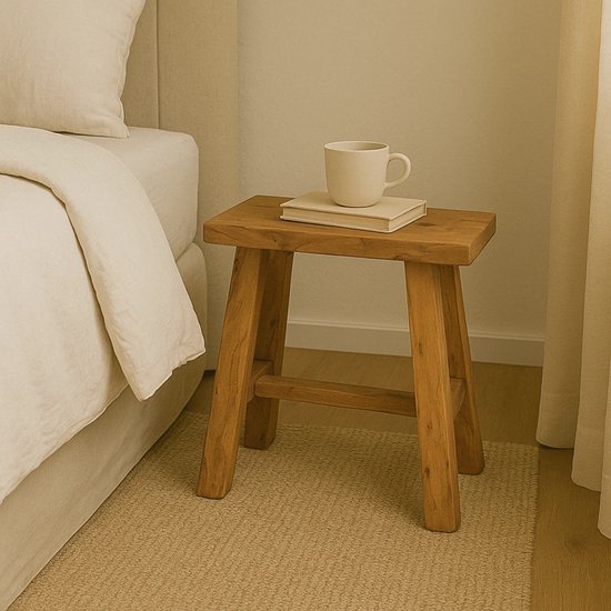 Een eenvoudig, houten krukje met een boek en mok op een nachtkastje, naast een bed.