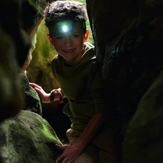 Jongen met een verlichte groene hoofdlamp in een rotsachtige omgeving, omhoog kijkend.