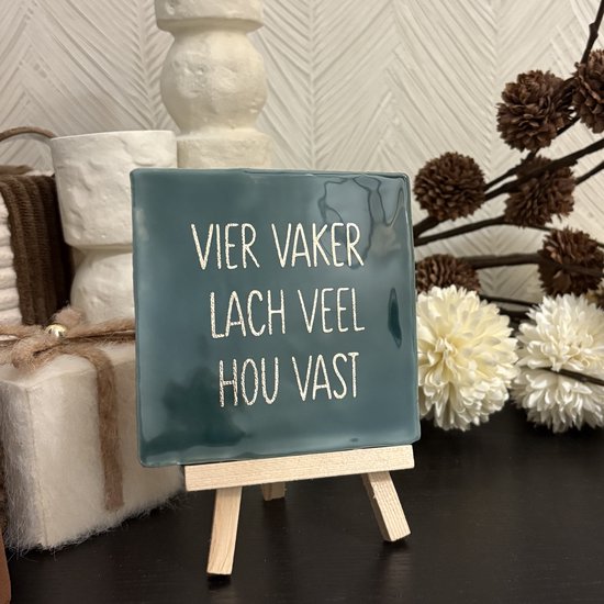 Donkergroen tegeltje met gouden tekst: 'Vier vaker, lach veel, hou vast'. Op een schildersezel.