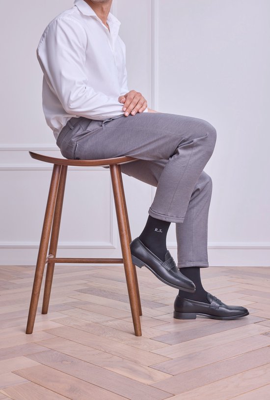 Homme assis sur un tabouret, portant un pantalon gris, des chaussettes noires avec initiales 'R.S.' et des mocassins noirs.