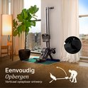 Zwarte roeitrainer verticaal opgeborgen naast een grote kamerplant en meubels.