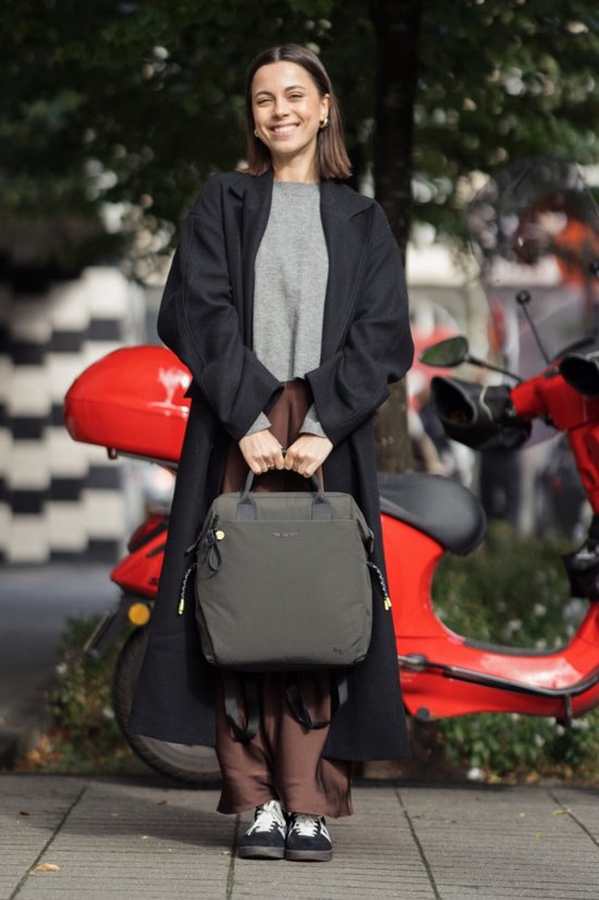 Vrouw met donkergroene rugzak, zwart jas, grijze trui en bruine broek, naast een scooter.