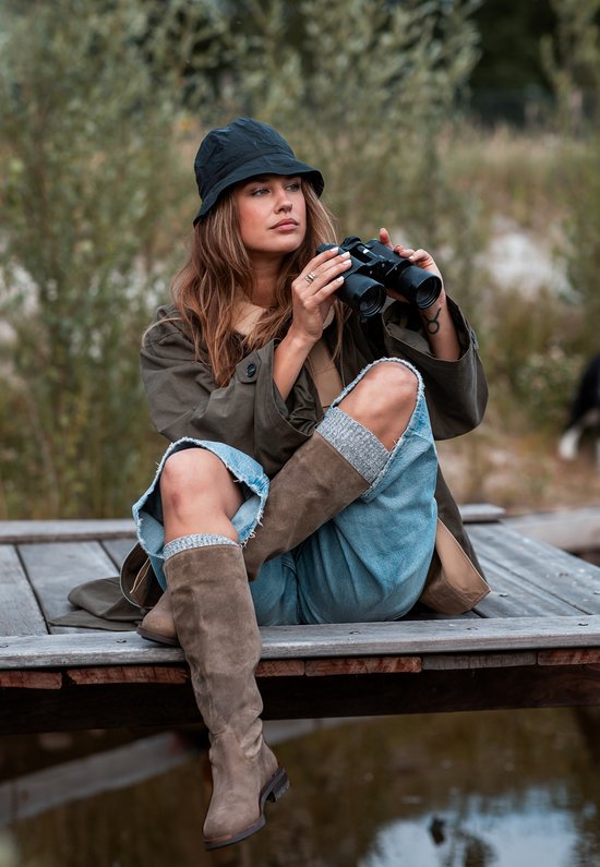 Model in bruine trenchcoat, blauwe jeans, bruine laarzen met grof profiel en zwarte hoed, met verrekijker.