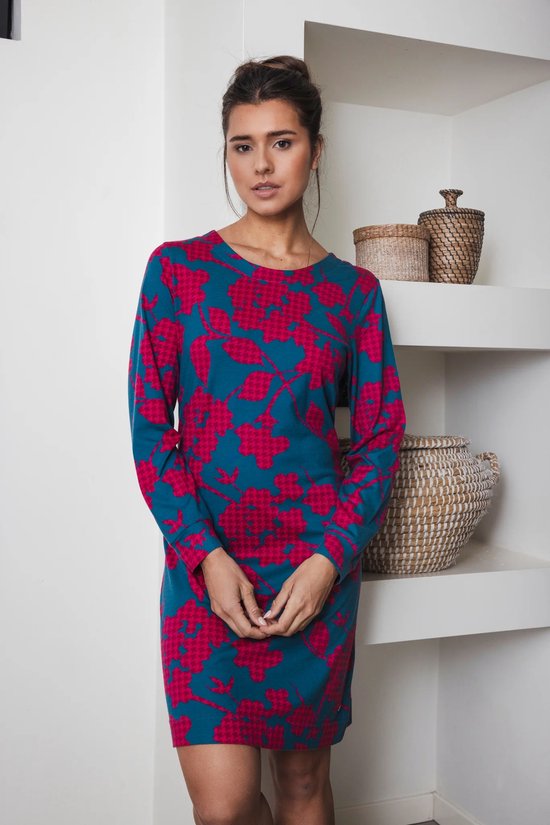 Vrouw met donkerbruin haar, draagt donkergroene jurk met lange mouwen en fuchsia bloemenprint, staat voor een kast.