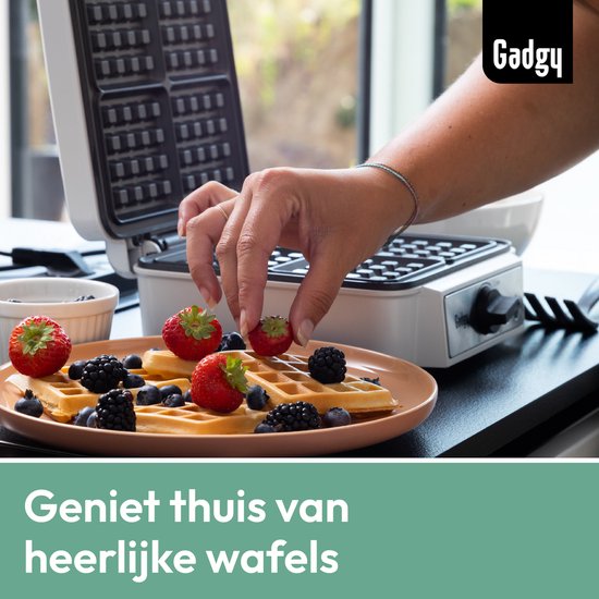 Hand versiert Brusselse wafels met aardbeien en bosbessen op een bord, met een wafelmaker op de achtergrond.