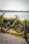 Lichtbruine elektrische fiets met zwarte details, bagagedrager, staand naast een hek en water.