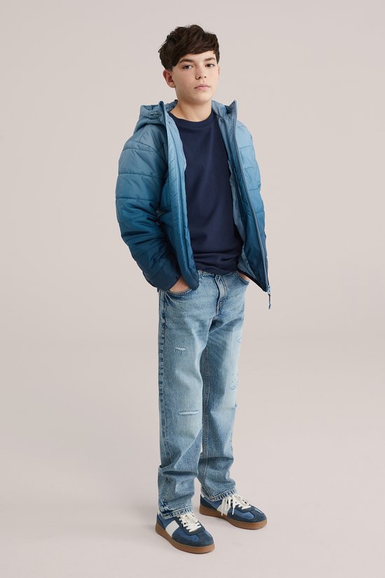 Jongen met de handen in de zakken, draagt een blauwe gewatteerde jas met spijkerbroek.
