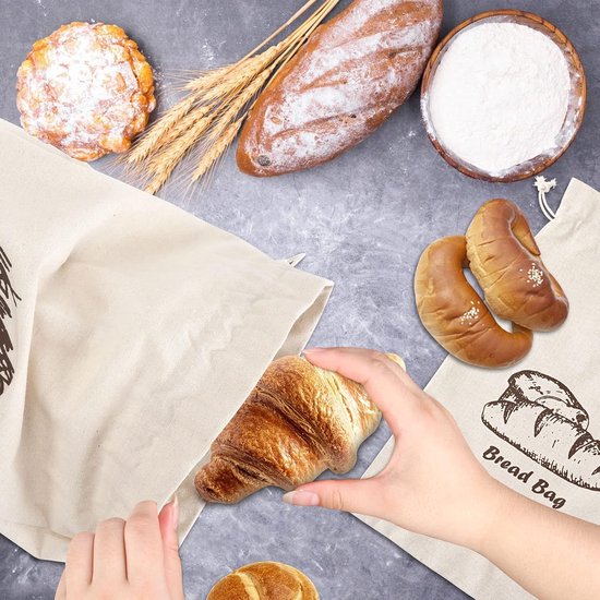 Een hand legt een croissant in een off-white canvas zak met broodprint, naast gebak en brood.