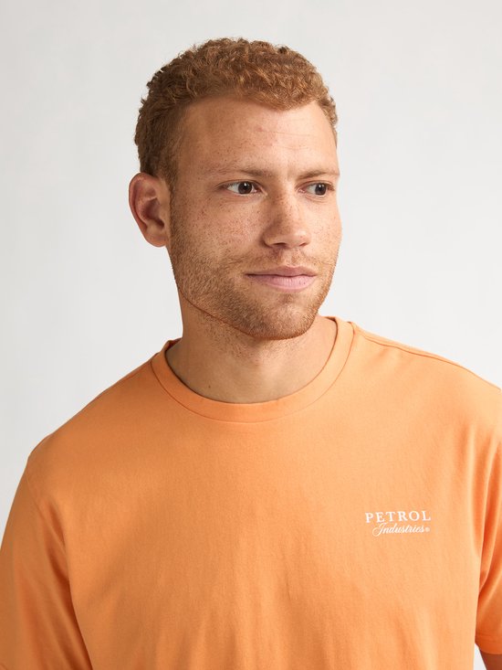 Close-up van een man met sproeten, die een oranje t-shirt met "PETROL Industries" op de borst draagt.
