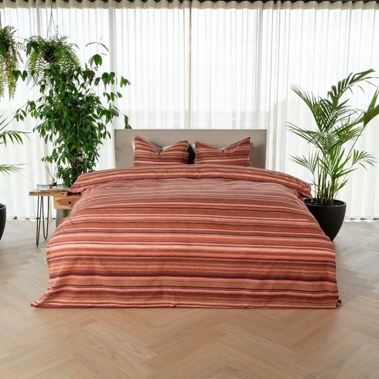 Bed opgemaakt met een dekbedovertrek en kussens met horizontale strepen in rood, bruin en roze tinten, in een slaapkamer met planten.
