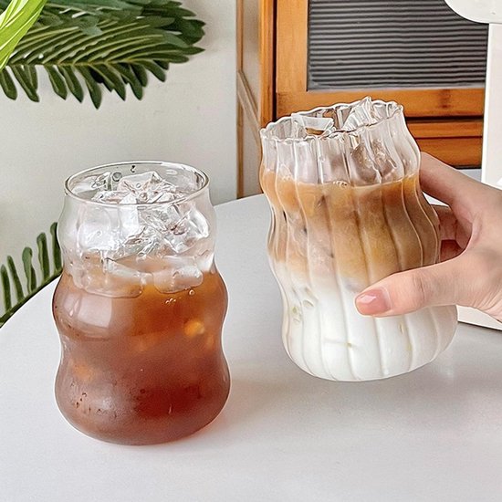 Twee gegolfde glazen bekers op een tafel. Eén gevuld met ijskoffie, de ander met melkdrank.