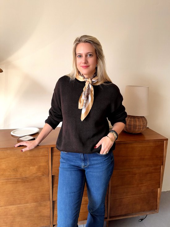 Model draagt een bruine trui, blauwe jeans en een crème sjaal met paisley-print om de nek.