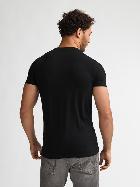 Man met handen in broekzakken, draagt zwarte T-shirt met korte mouwen en grijze jeans.