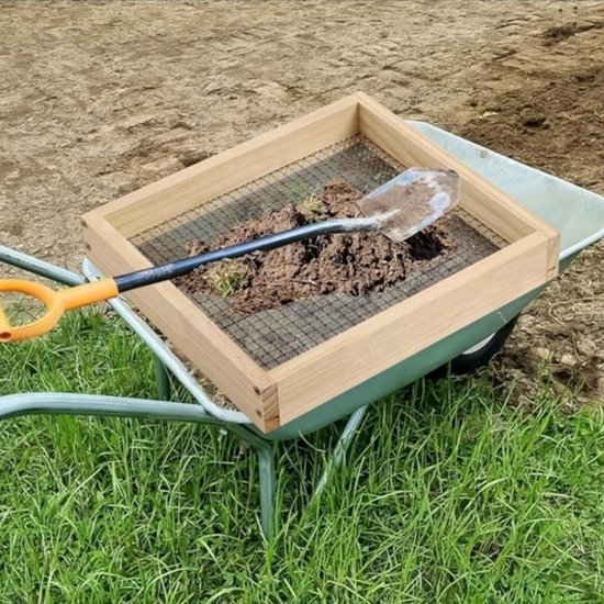 Een houten zeef met metalen gaas staat op een kruiwagen met aarde. Een schep ligt op de zeef.
