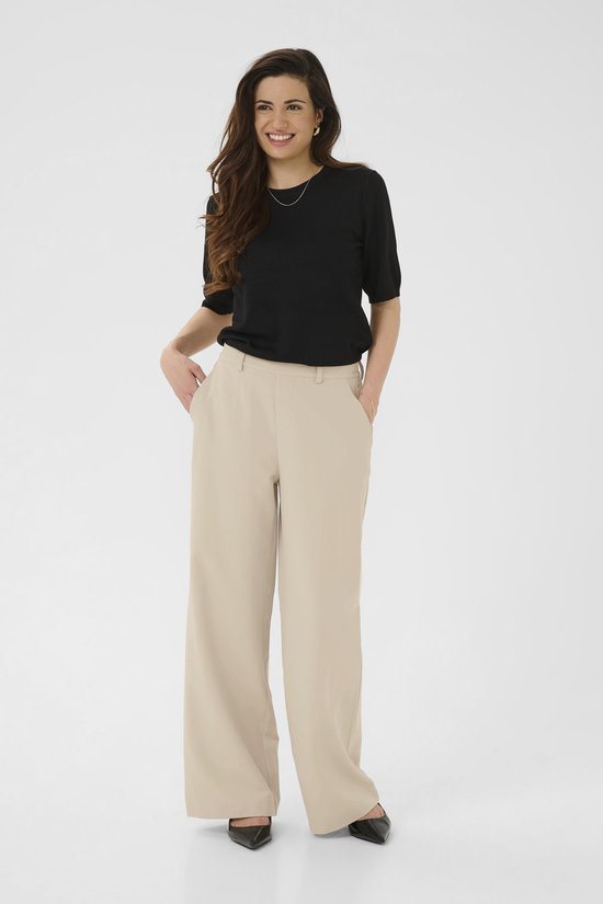 Model in een zwarte top en een beige wijde broek, staand met handen in de zakken.
