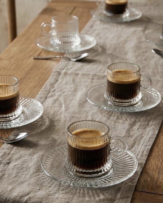 Six tasses à expresso en verre avec soucoupes, remplies de café, sur une table.