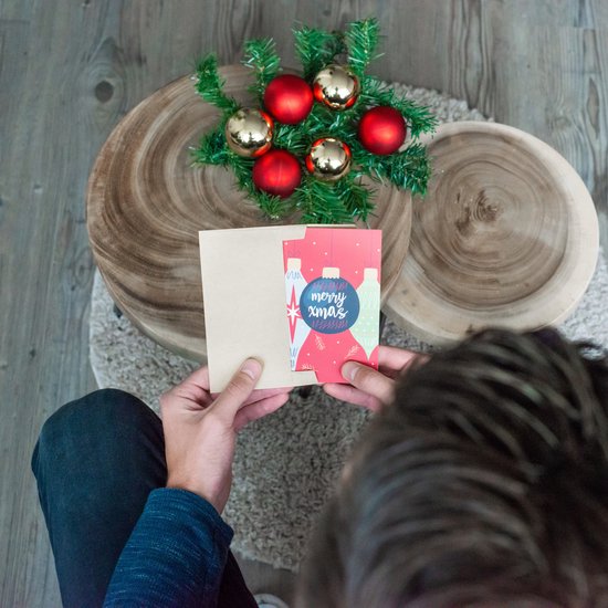 Man houdt kerstkaart met kerstballen en tekst 'merry xmas' vast. Op de achtergrond kerstdecoratie en houten tafels.