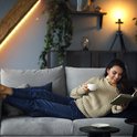 Vrouw in trui en spijkerbroek leest een boek op de bank met een kop koffie, verlicht door een gele led-strip.