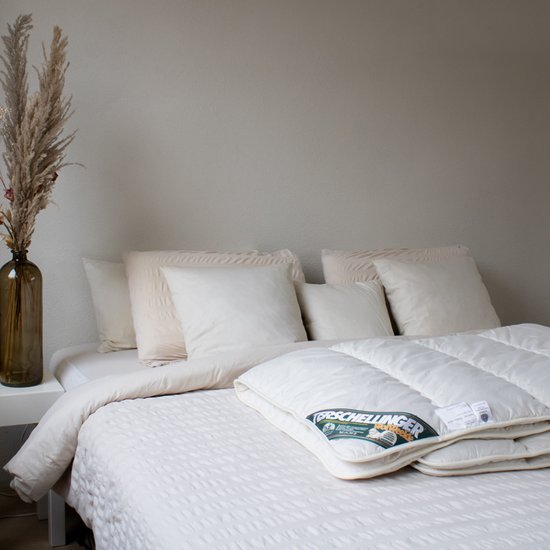 Wit gestikt dekbed op een bed met beige kussens en een vaas met droogbloemen.