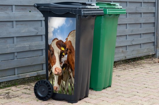 Poubelle noire avec un autocollant de vaches marron et blanches avec des étiquettes jaunes, à côté d'une poubelle verte.