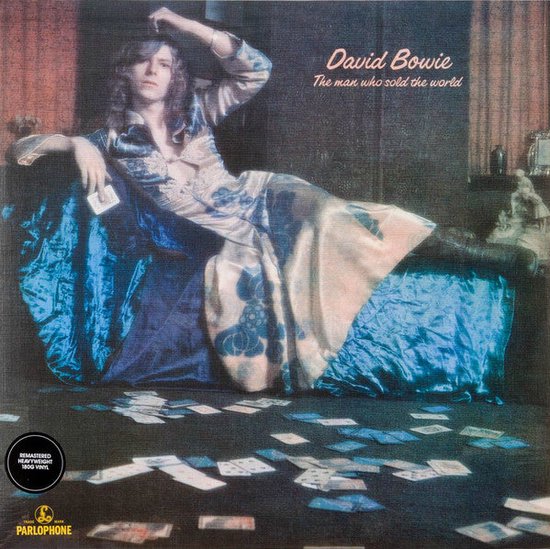 David Bowie op een bank in een bloemenjurk, met speelkaarten verspreid over de vloer. 'David Bowie The man who sold the world' in roze tekst.