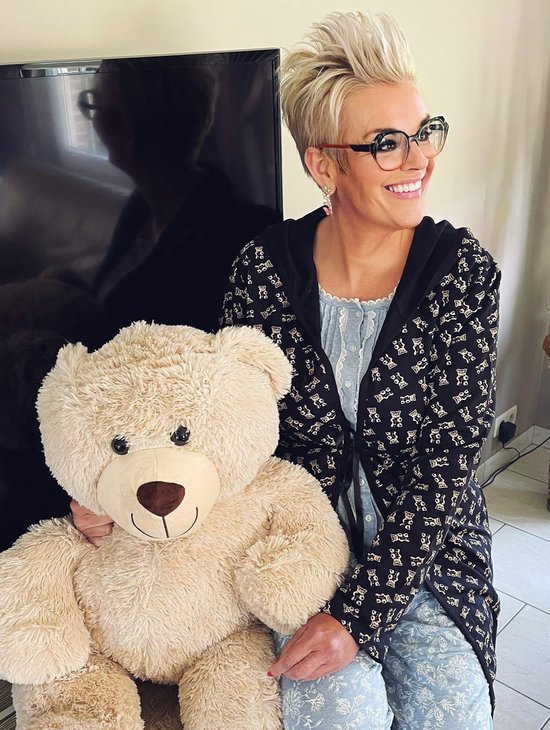 Une femme aux cheveux blonds et portant des lunettes, tient un gros ours en peluche et porte un peignoir noir à imprimé ours en peluche.