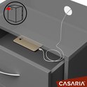 Close-up van een open lade van een grijs nachtkastje, waarin een smartphone oplaadt via een kabel. Een ronde opening voor kabels is zichtbaar.