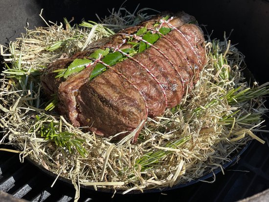 Geroosterde rollade met kruiden en touw, liggend op hooi en rozemarijn in een gietijzeren pan.