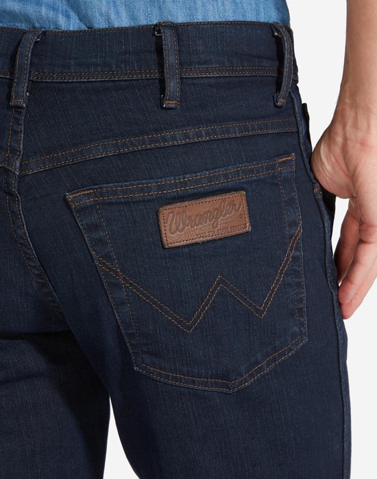 Close-up van de achterzak van een donkerblauwe Wrangler jeans met iconische stiksels en een bruine merkpatch.