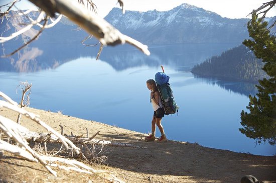 Femme portant un grand sac à dos marche sur un sentier de montagne, avec un lac bleu et des montagnes en arrière-plan.