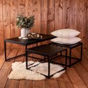 Ensemble de tables basses noires avec une grande et deux petites tables sur un plancher en bois, décoré d'un vase et de coussins.