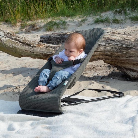 Baby in een groen wafelstof wipstoeltje op een strand, naast een boomstam.