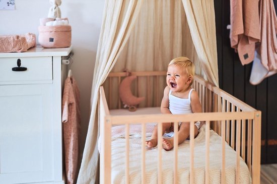 Een lachende baby met een wit hemdje zit in een ledikant onder een lichtbeige hemeltje.