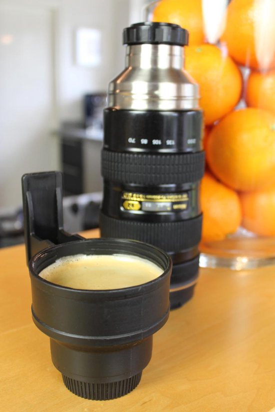 Zwarte thermosfles in de vorm van een cameralens, met een mok en sinaasappels op de achtergrond.