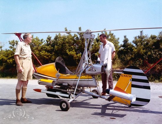 Twee mannen naast een gele en zilverkleurige gyrocopter met een oranje staartvin.