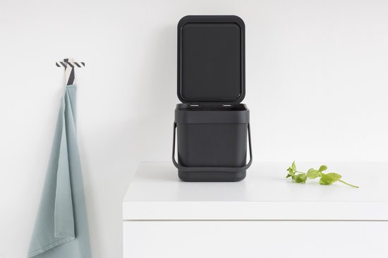Boîte à compost Brabantia gris foncé avec couvercle ouvert sur un plan de travail blanc.