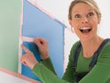 Vrolijke vrouw met blond haar brengt roze schilderstape aan op een blauw vlak op een muur.