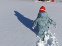 Kind in een lichtgroen regenpak met visgraatpatroon, oranje muts en groene laarzen, lopend in de sneeuw.