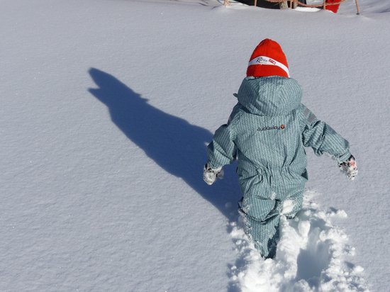 Kind in een lichtgroen regenpak met visgraatpatroon, oranje muts en groene laarzen, lopend in de sneeuw.